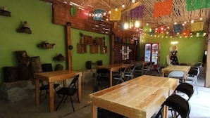 A rustic interior with wooden tables and chairs, a green wall with shelves, and colorful hanging decorations. - Hotel Los Ciruelos (Oaxaca)