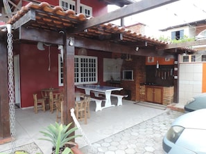 Une terrasse extérieure dotée d’une pergola en bois, d’une table à manger avec des chaises, d’un barbecue et d’une voiture garée à proximité.