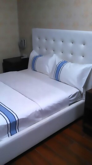 A neatly made bed with white bedding and blue stripes, a tufted headboard, and a nightstand with a lamp. - Hotel Spa Bella Mar (Santo Domingo)