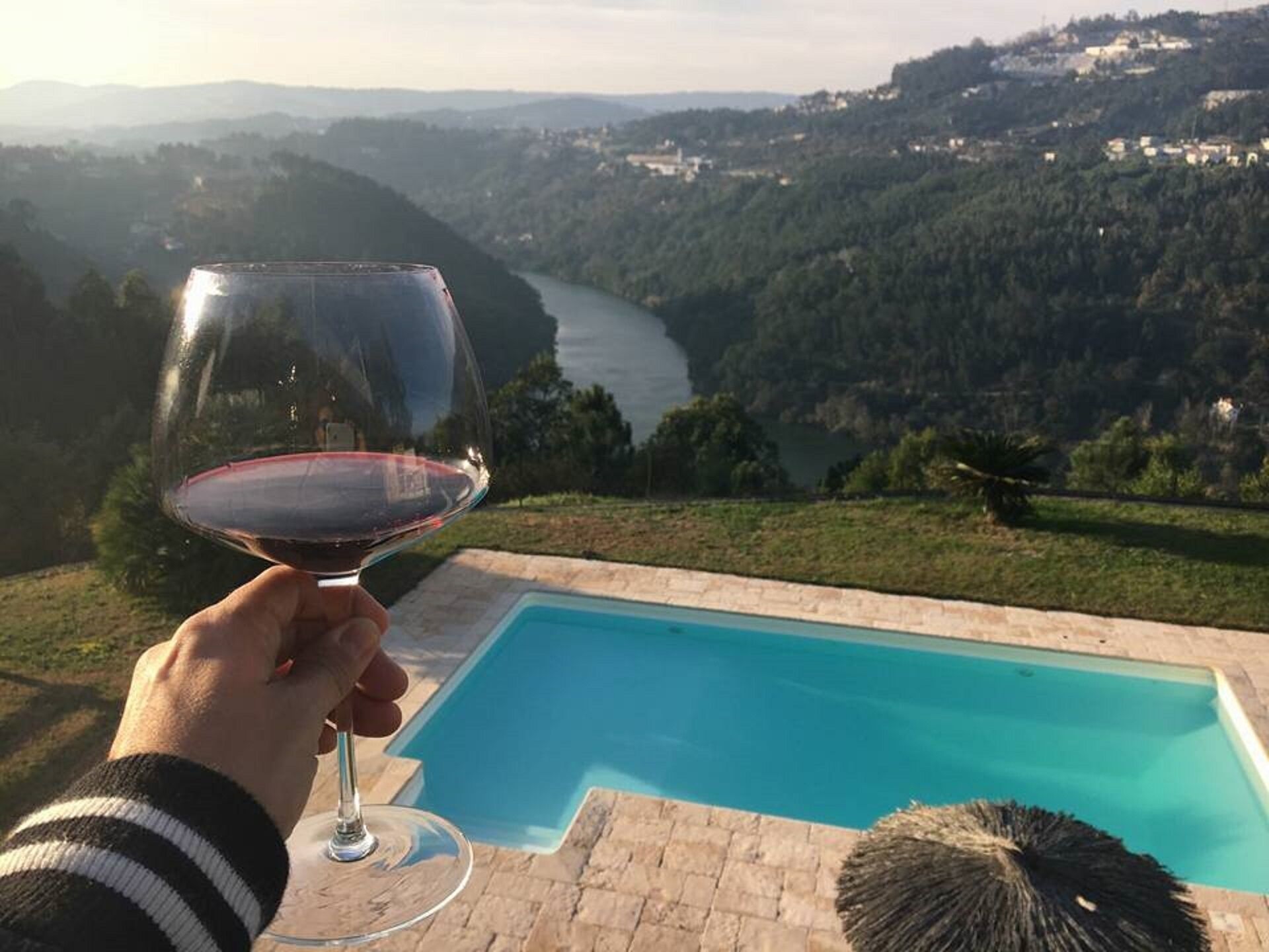 Een hand die een wijnglas vasthoudt, met zicht op een zwembad en een rivier op de achtergrond.