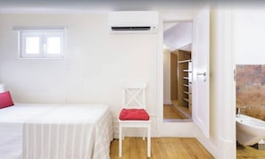 A bedroom with a bed, a chair, an air conditioning unit, a window, and a bathroom with a bathtub.