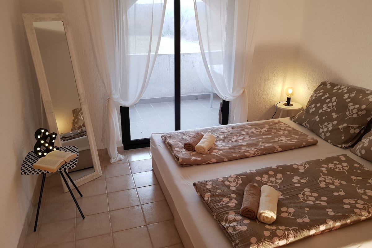 Een slaapkamer met een bed, een nachtkastje met lamp, een spiegel en een raam met dunne gordijnen.