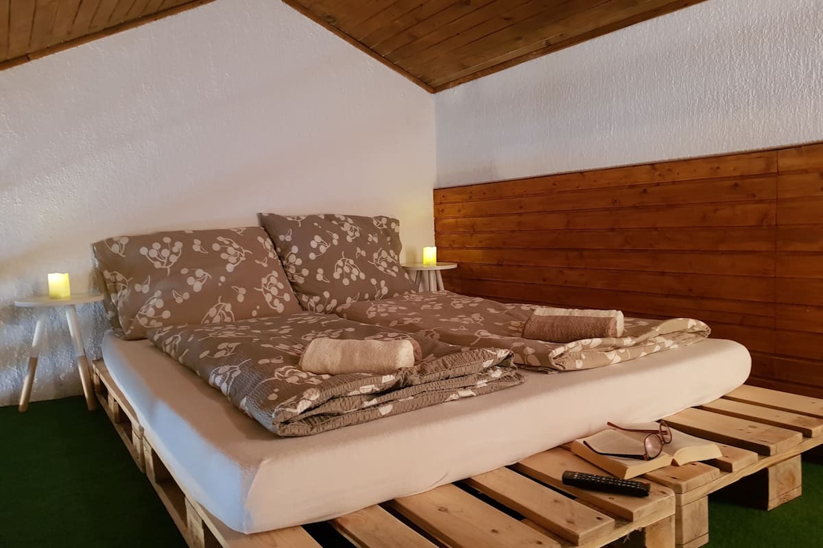 Een bed met een bloempatroon sprei en kussens, een nachtkastje met een kaars, en een houten bankje met een afstandsbediening en brillen.