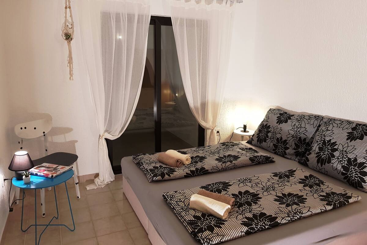 Een slaapkamer met een bed, een nachtkastje met een lamp en een raam met dunne gordijnen.