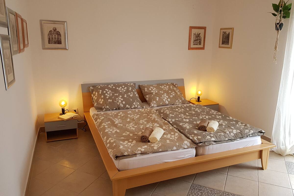 Een tweepersoonsbed met bloemenbedrukking, twee nachtkastjes met lampen, en ingelijste foto's aan de muur.
