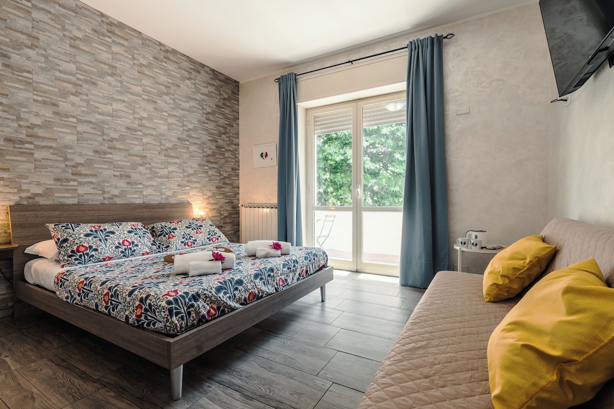 A modern bedroom with a bed, a sofa, a window with curtains, a radiator, and a wall with a textured pattern.