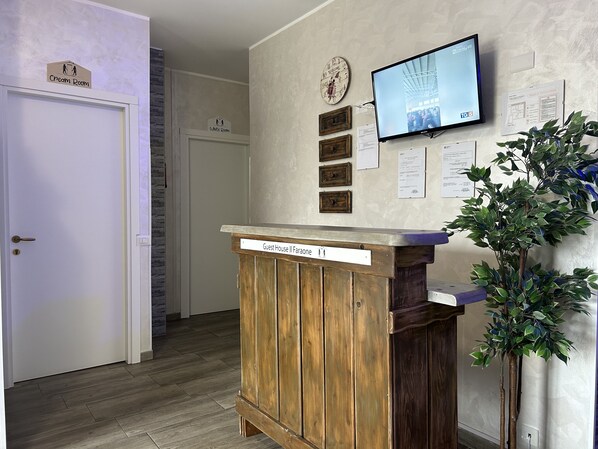A reception area with a wooden counter labeled "Guest House II Faunae," a television mounted on the wall, and a clock with a "Welcome" sign.
