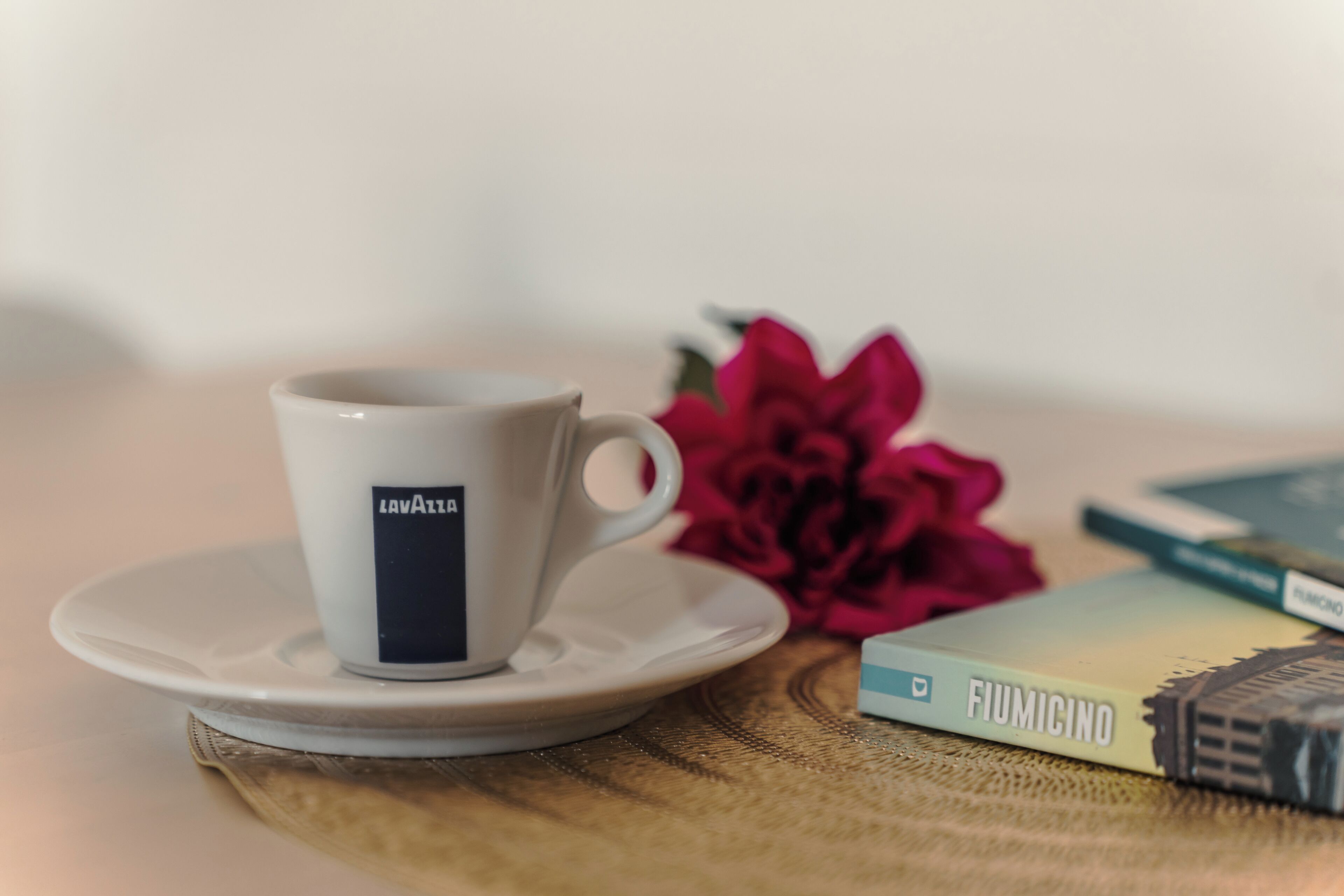 Een witte kop met een zwart etiket op een schoteltje, een rode bloem en een boek met de titel 'FIUMICINO' op een tafel.