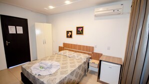 A hotel room with a bed, a small refrigerator, a wardrobe, and an air conditioning unit. - Pousada Juliane (Penha)
