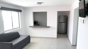 Una sala moderna con un sofá gris, una estantería empotrada, una zona de cocina con refrigerador y un televisor montado en la pared.