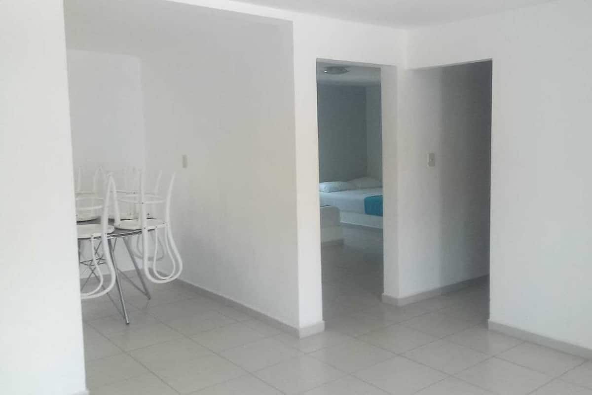 A spacious, well-lit room with white walls and a tiled floor, featuring a dining area with white chairs and a visible bed in the background.