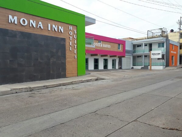 A street view of a hotel named 'Mona Inn' with 'Hotel & Suites' signage, featuring a modern facade with a green accent and a visible balcony.