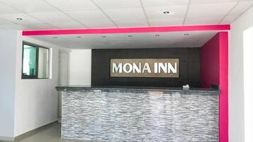 Uma área de recepção com o letreiro 'MONA INN', balcão com textura de pedra e uma parede de destaque rosa vibrante.