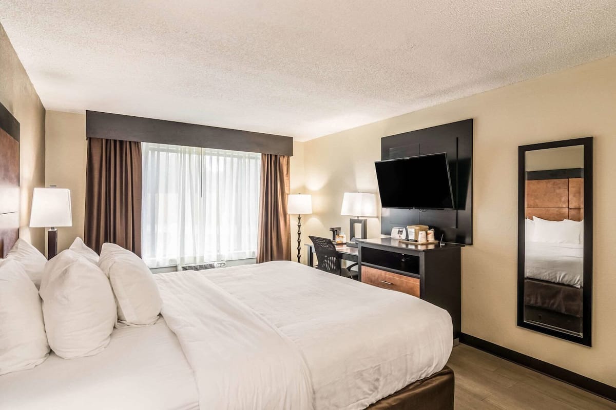 A hotel room with a large bed, a desk with a flat-screen TV, a mirror, and a window with curtains.