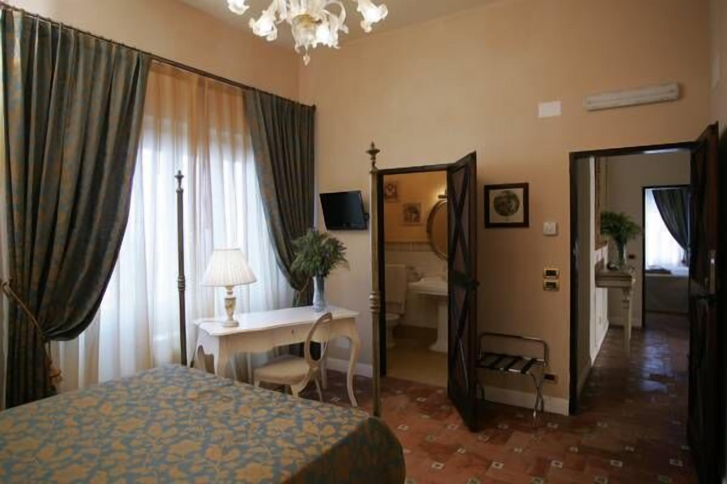A hotel room with a bed, a desk, a chair, a lamp, a potted plant, and a bathroom visible through an open door.