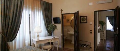 A hotel room with a bed, a desk, a chair, a lamp, a potted plant, and a bathroom visible through an open door.