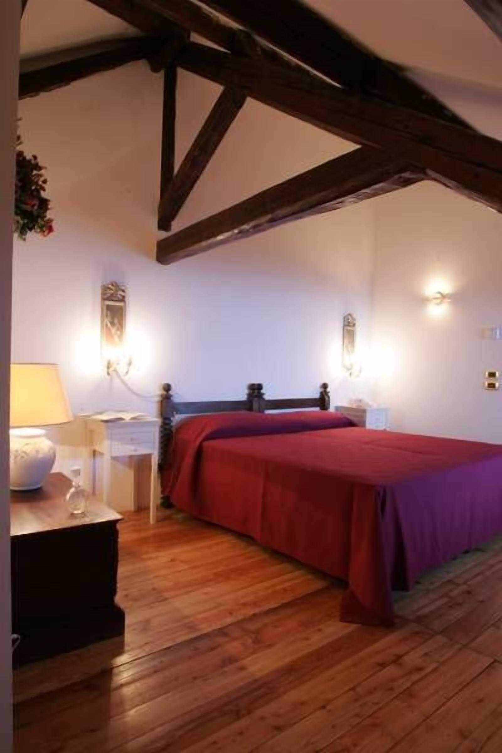 Une chambre avec un plancher en bois, un lit recouvert d’une courtepointe rouge, une table de chevet avec une lampe et des appliques murales.