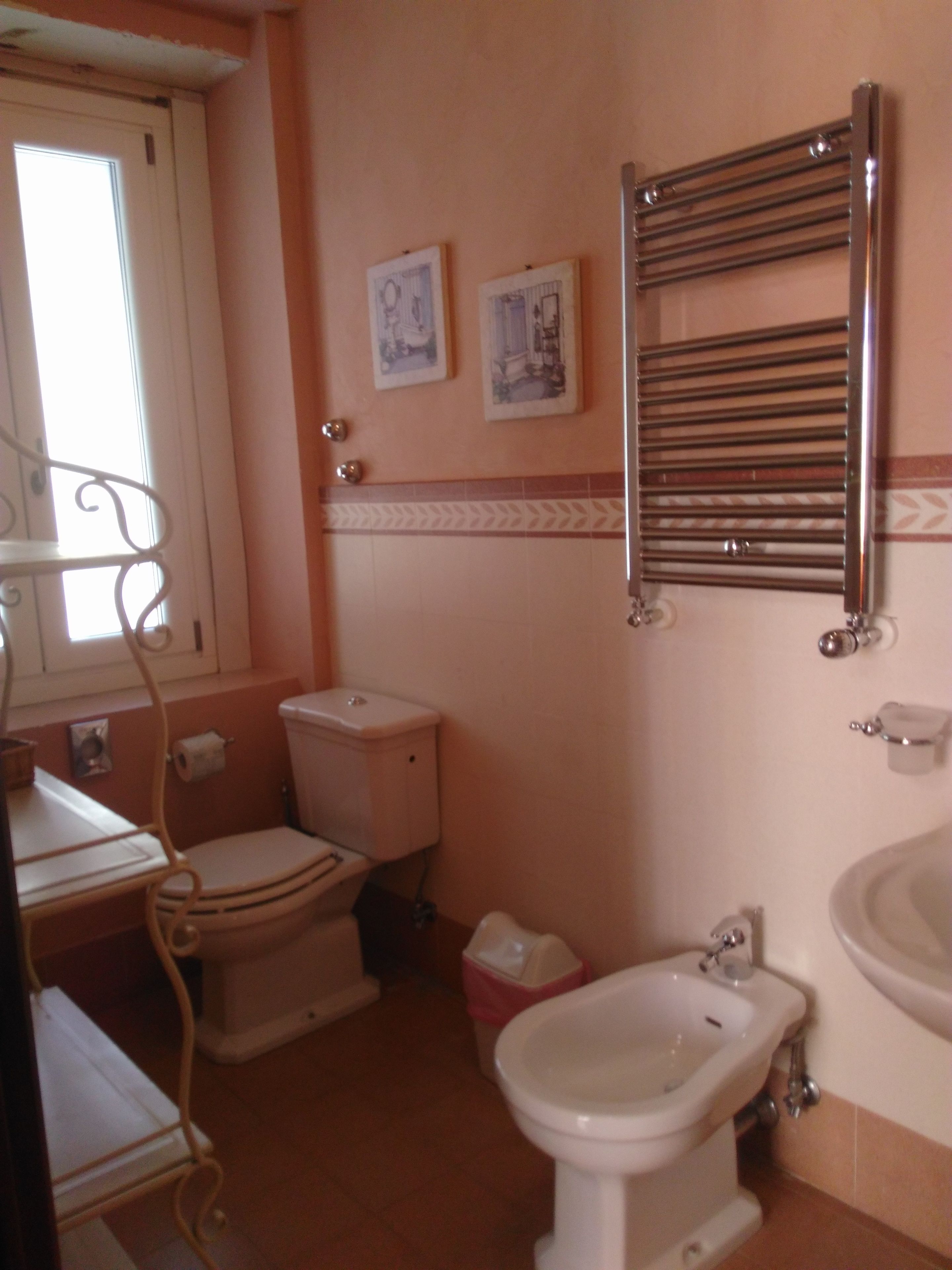 Un baño con inodoro, bidé y lavabo. Hay una ventana con cortinas, un perchero y dos cuadros en la pared.