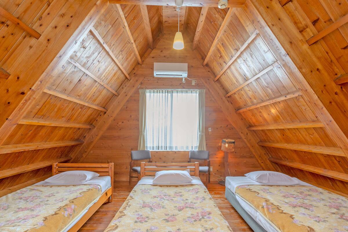A cozy attic bedroom with wooden walls and a sloped ceiling. There are two beds with floral bedspreads and a small table with chairs.