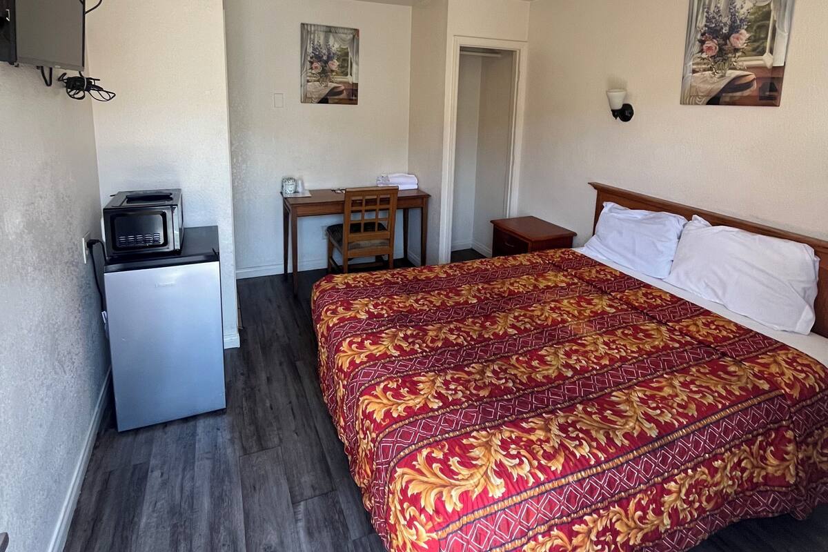 A bedroom with a bed, a desk, a chair, a small refrigerator, and a television on the wall.