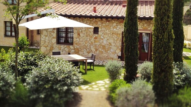A stone building with a tiled roof, a patio with a table and chairs, and a well-maintained garden with shrubs and trees.