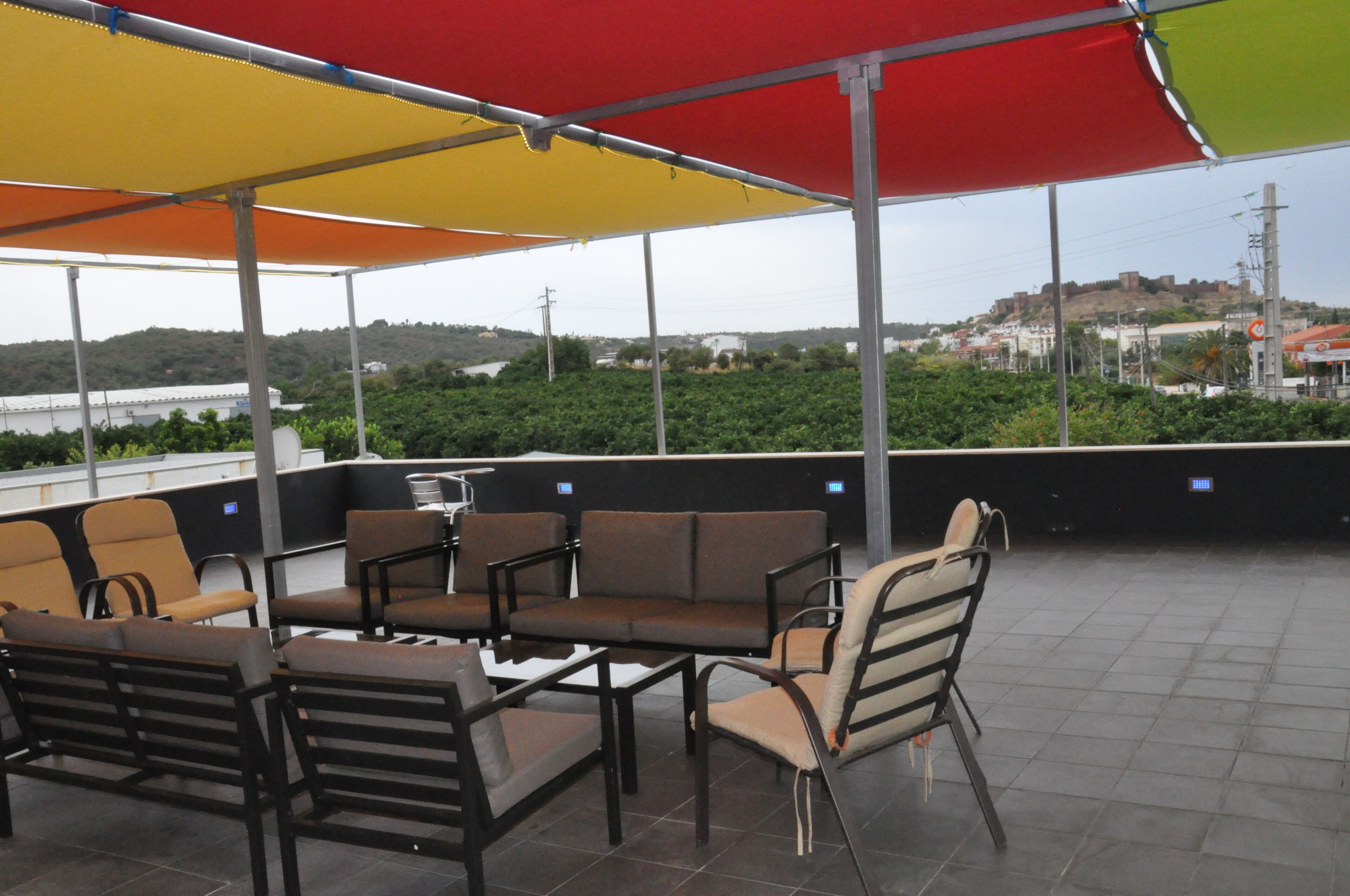 An outdoor seating area with multiple chairs and tables, a large umbrella providing shade, and a view of a distant hill with buildings.