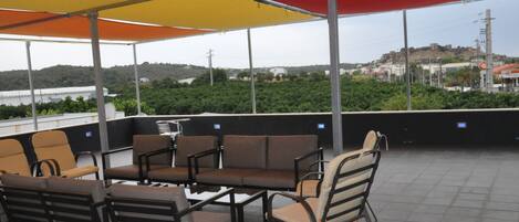 An outdoor seating area with multiple chairs and tables, a large umbrella providing shade, and a view of a distant hill with buildings.