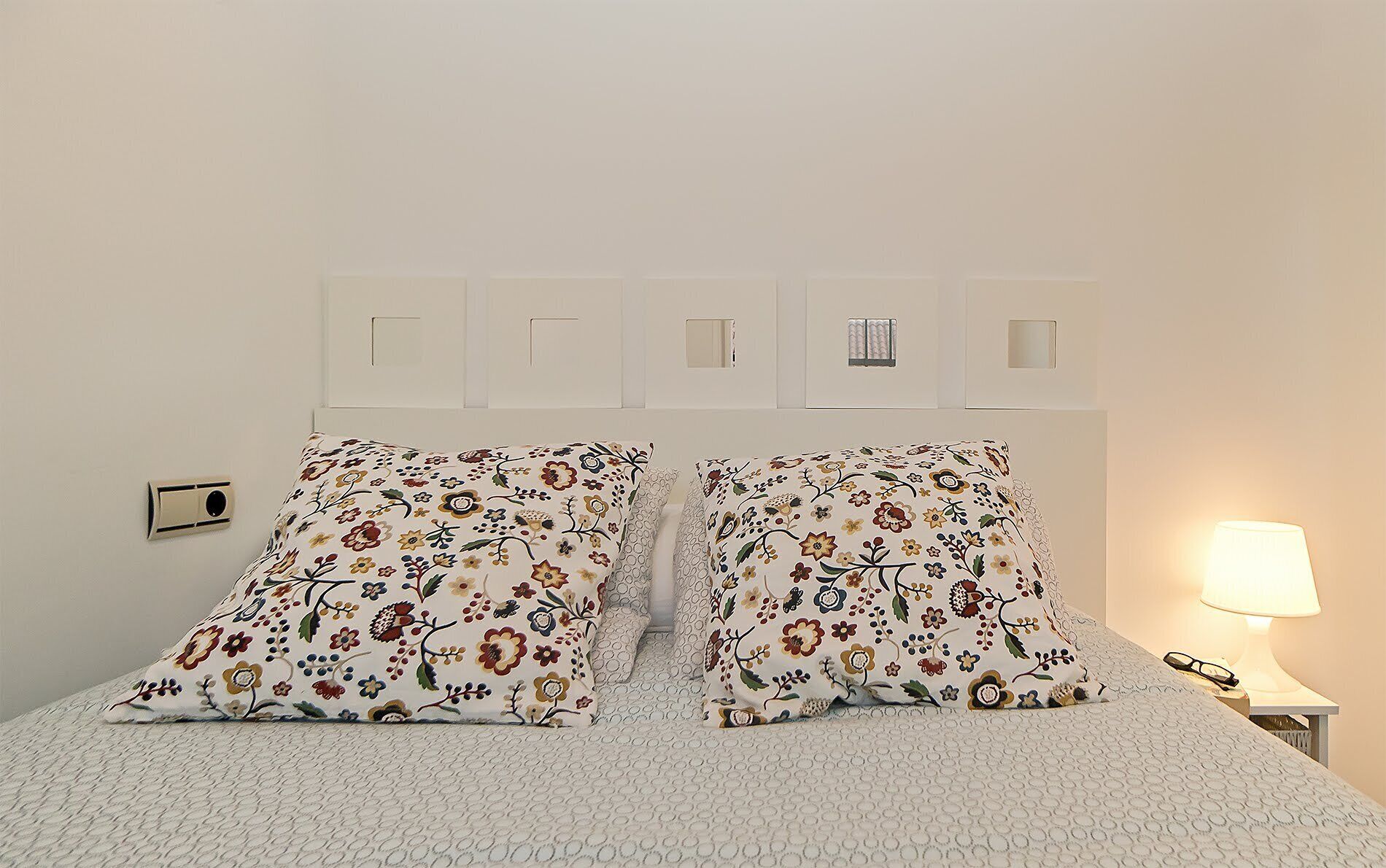 Una cama con almohadas estampadas con motivos florales, un cabecero blanco con interruptores de luz y una lámpara de noche.