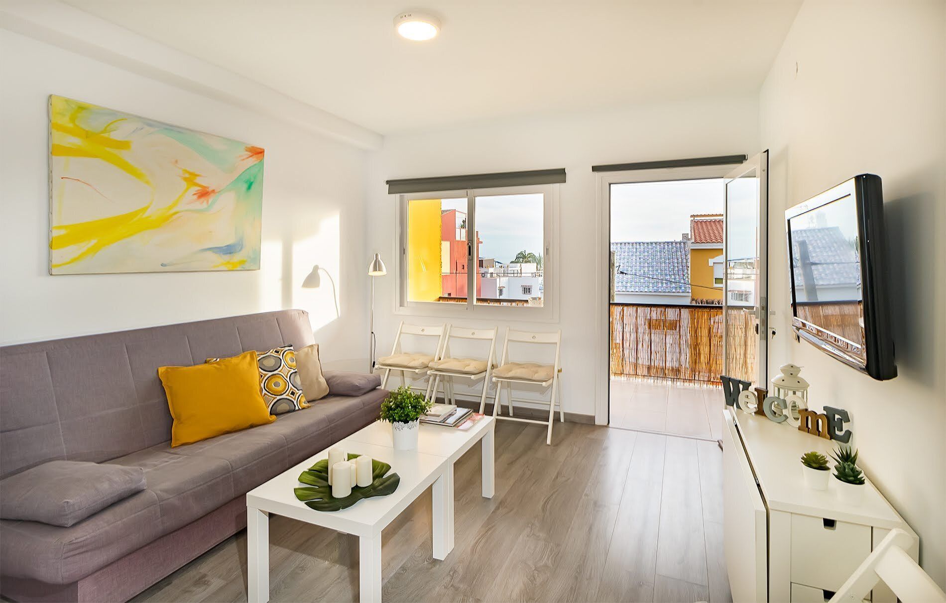 A modern living room with a grey sofa, white coffee table, and a painting on the wall. There are two windows with views of buildings and a balcony with a wooden fence.
