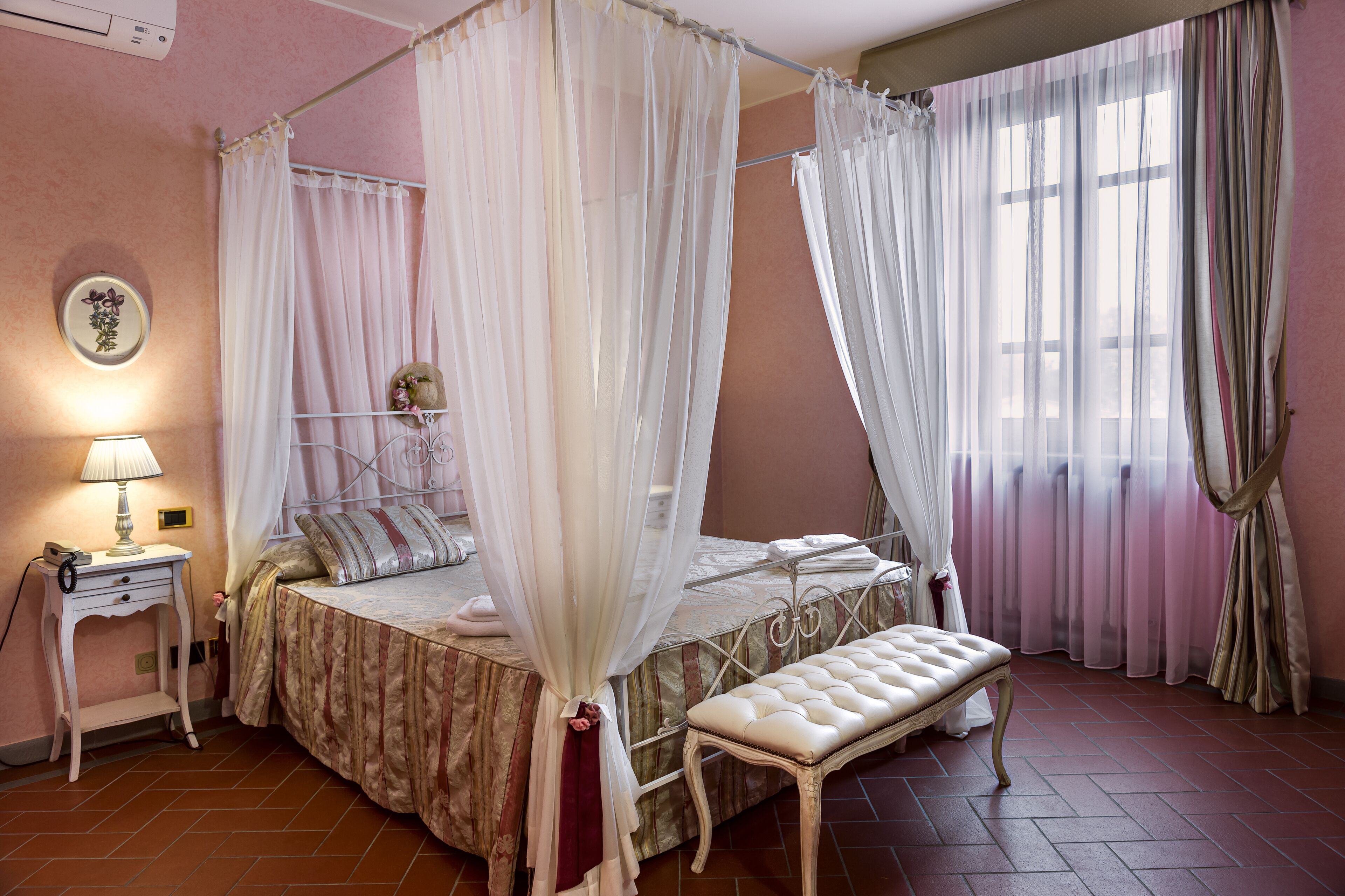 A bedroom with a four-poster bed, a bedside table with a lamp, a bench, and a window with sheer curtains.