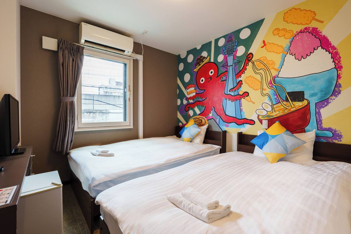 A hotel room with two beds, a desk with a computer, and a colorful mural on the wall.