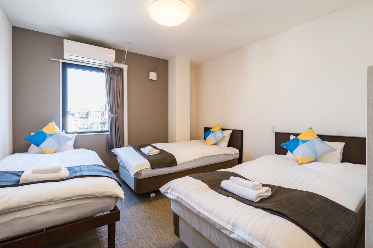 A hotel room with three beds, a window with curtains, an air conditioning unit, and a city view.