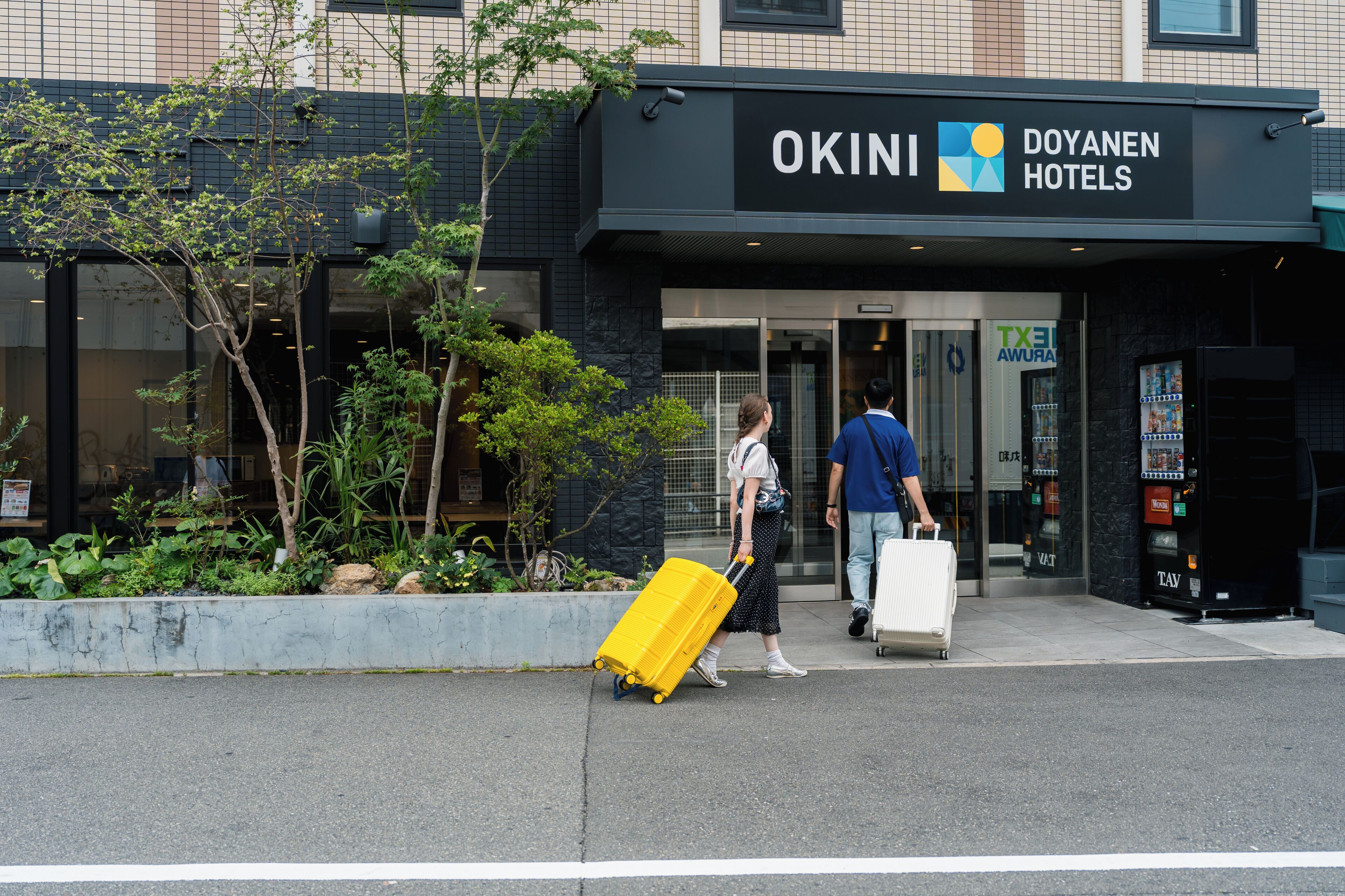 Egy szálloda bejárata, amelyen az „OKINI Doyan Hotels” feliratú tábla látható, és egy személy húz egy sárga színű bőröndöt.