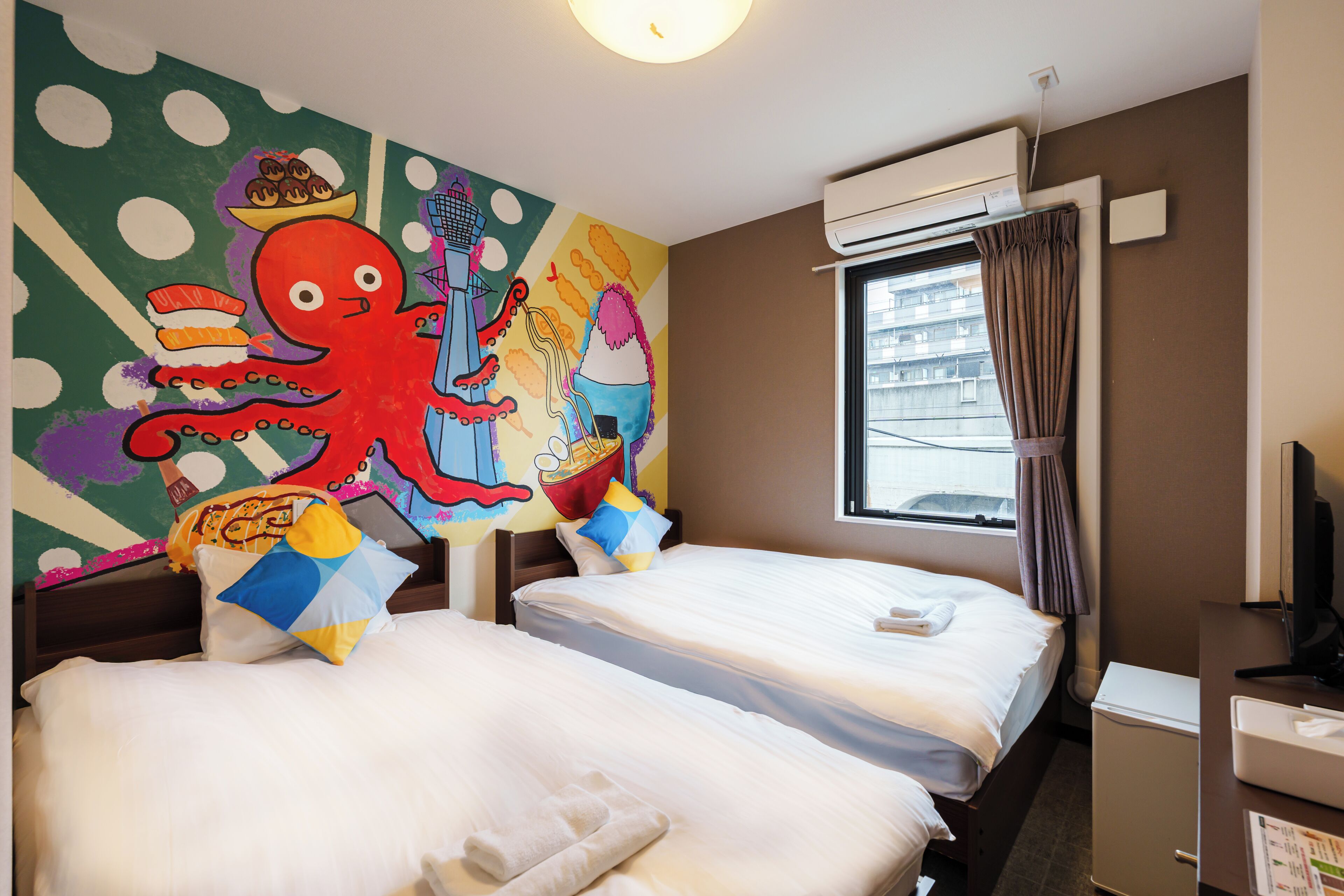 Habitación de hotel con dos camas, un mural grande y colorido en la pared, un equipo de aire acondicionado, una ventana con cortinas y un escritorio con una computadora.
