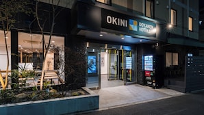 A hotel entrance with a sign for 'OKINI Doyaneni Hotels,' a glass door entrance, and a vending machine outside. - OKINI - DOYANEN HOTELS (Osaka)