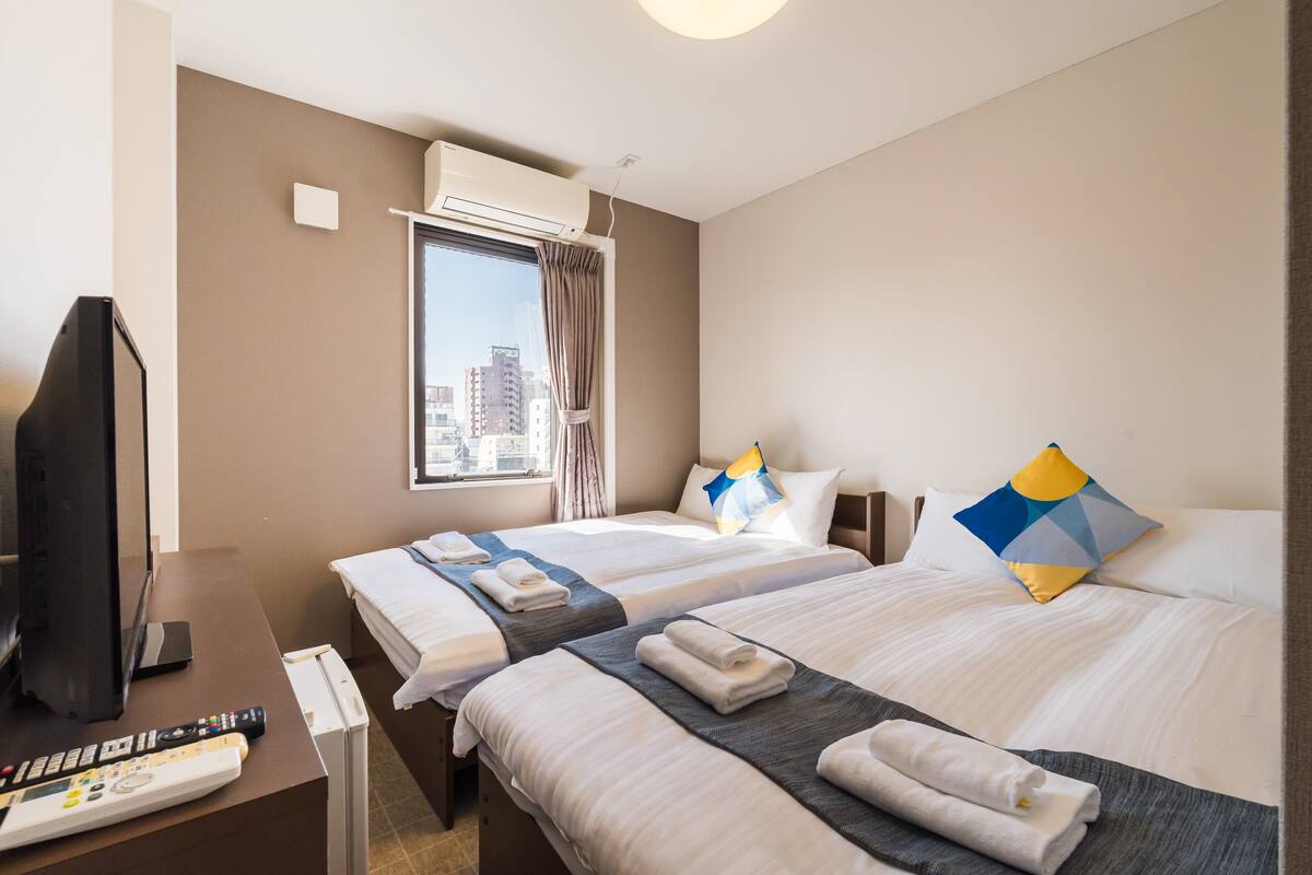 A hotel room with two beds, a TV, a desk with a remote control, and an air conditioning unit.