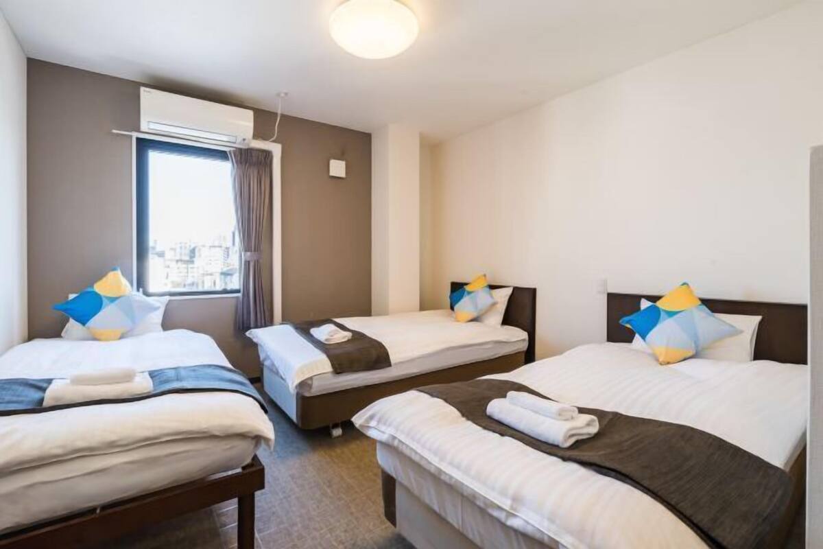 A hotel room with three beds, a window with curtains, an air conditioning unit, and a city view.