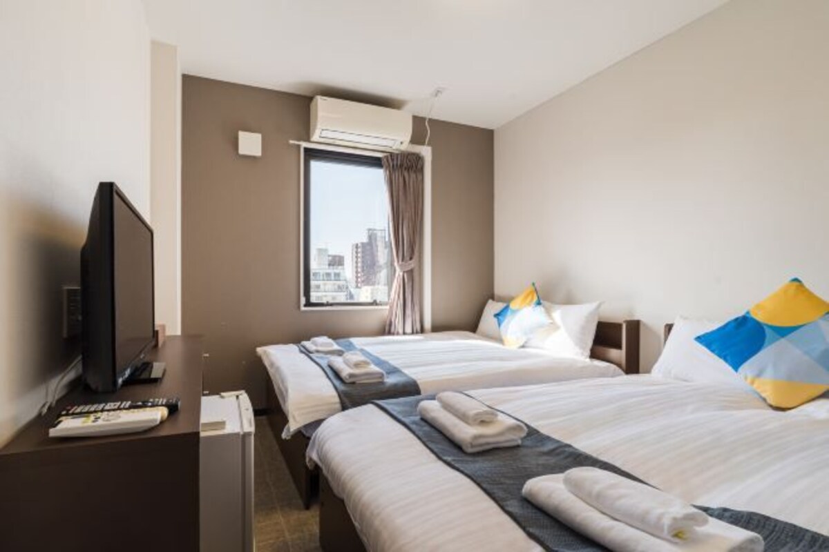 A hotel room with two beds, a TV, a desk, and an air conditioning unit.