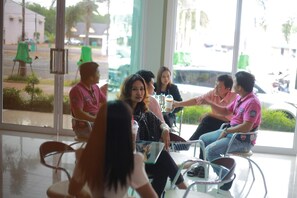 A group of people seated around a table inside a building with large windows, some chairs are green, and there is a person standing near the windows.