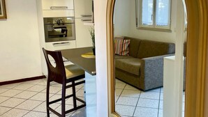 Une cuisine compacte dotée d’appareils électroménagers modernes, une table à manger avec une chaise, et un canapé gris dans un coin salon avec une fenêtre.
