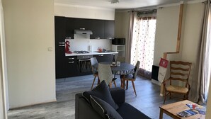 En moderne stue med en sort sofa, et træbord og et spiseområde med et rundt bord og stole. Et køkken med sorte skabe og en gasblus ses i baggrunden.