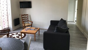 En stue med en sort sofa, en træstol, et sofabord med magasiner, et fjernsyn monteret på væggen og en gulvtæppe med et mønster.