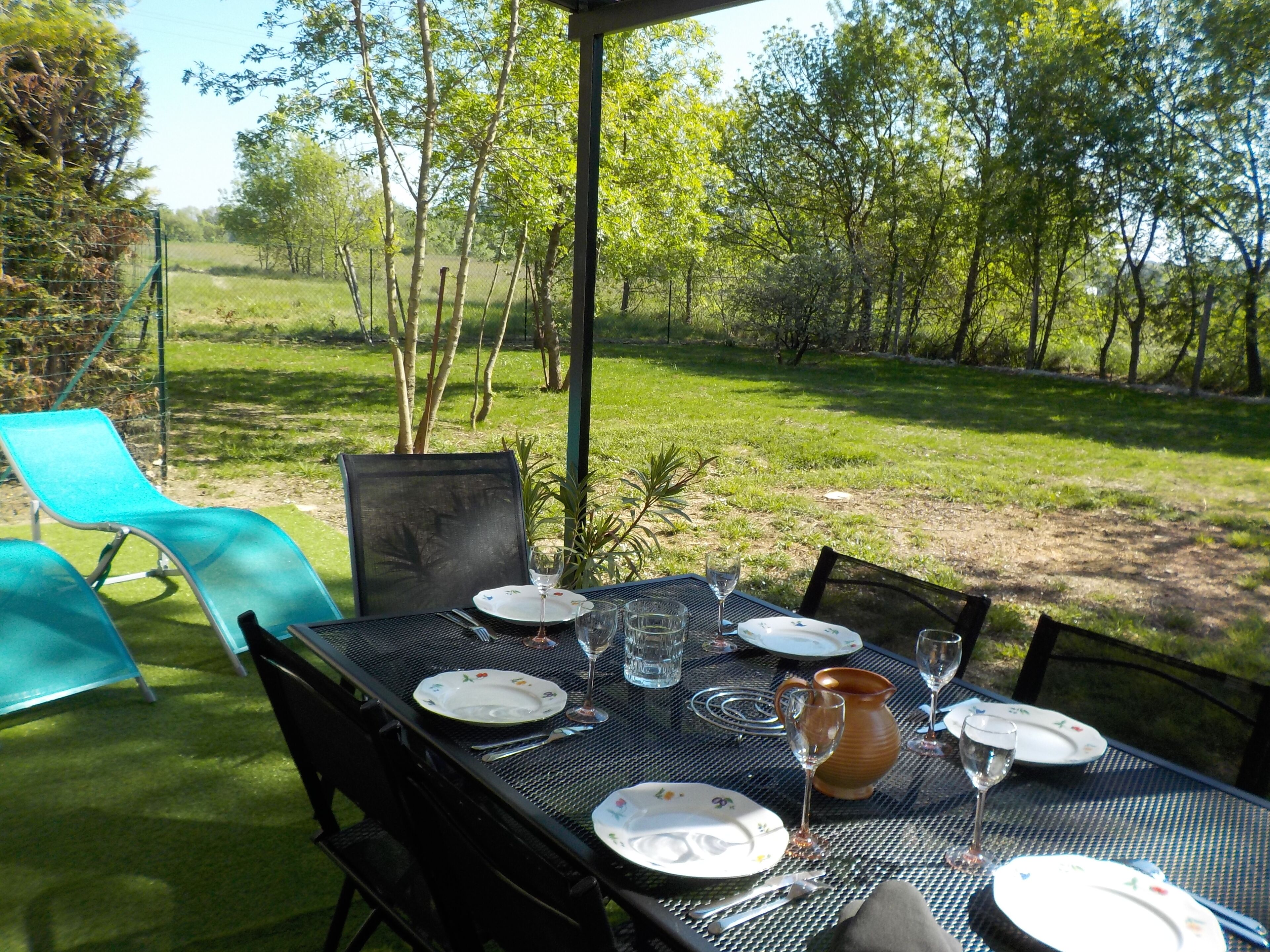 Un espace de restauration en plein air avec une table dressée pour quatre personnes, entourée de chaises, dans un cadre de jardin luxuriant.