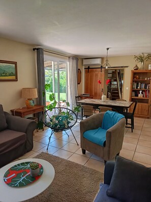 A living room with a sofa, armchair, dining table, and a view of the outdoors through a glass door.