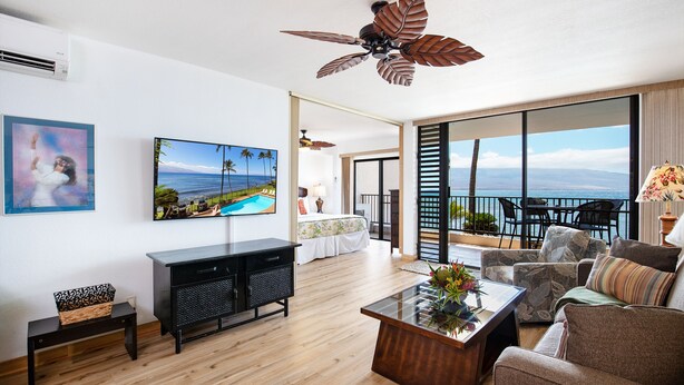 Un salón moderno con un televisor de pantalla plana grande, un ventilador de techo y un balcón con vistas al océano.