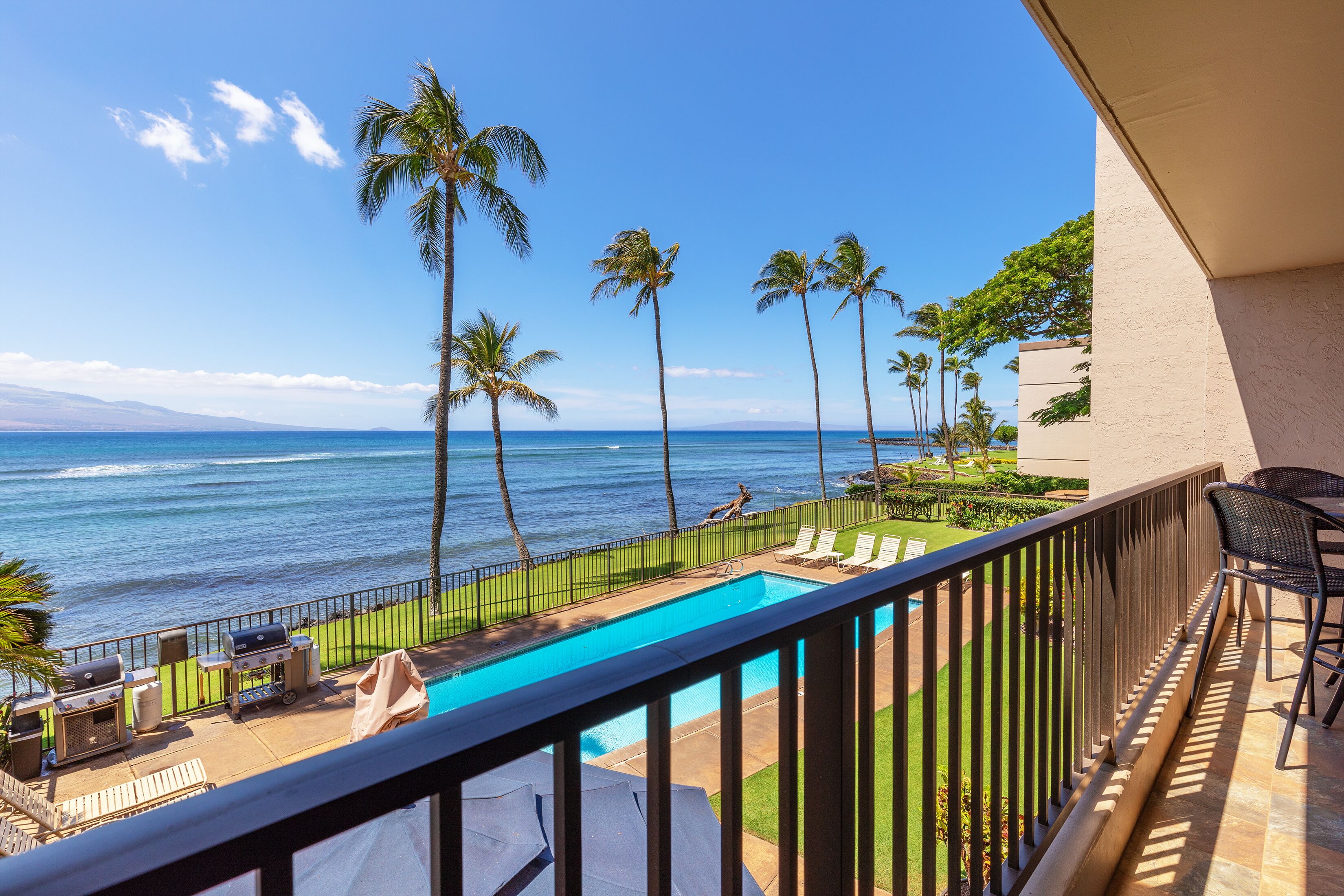 A balcony with a view of the ocean, a swimming pool, and lounge chairs.