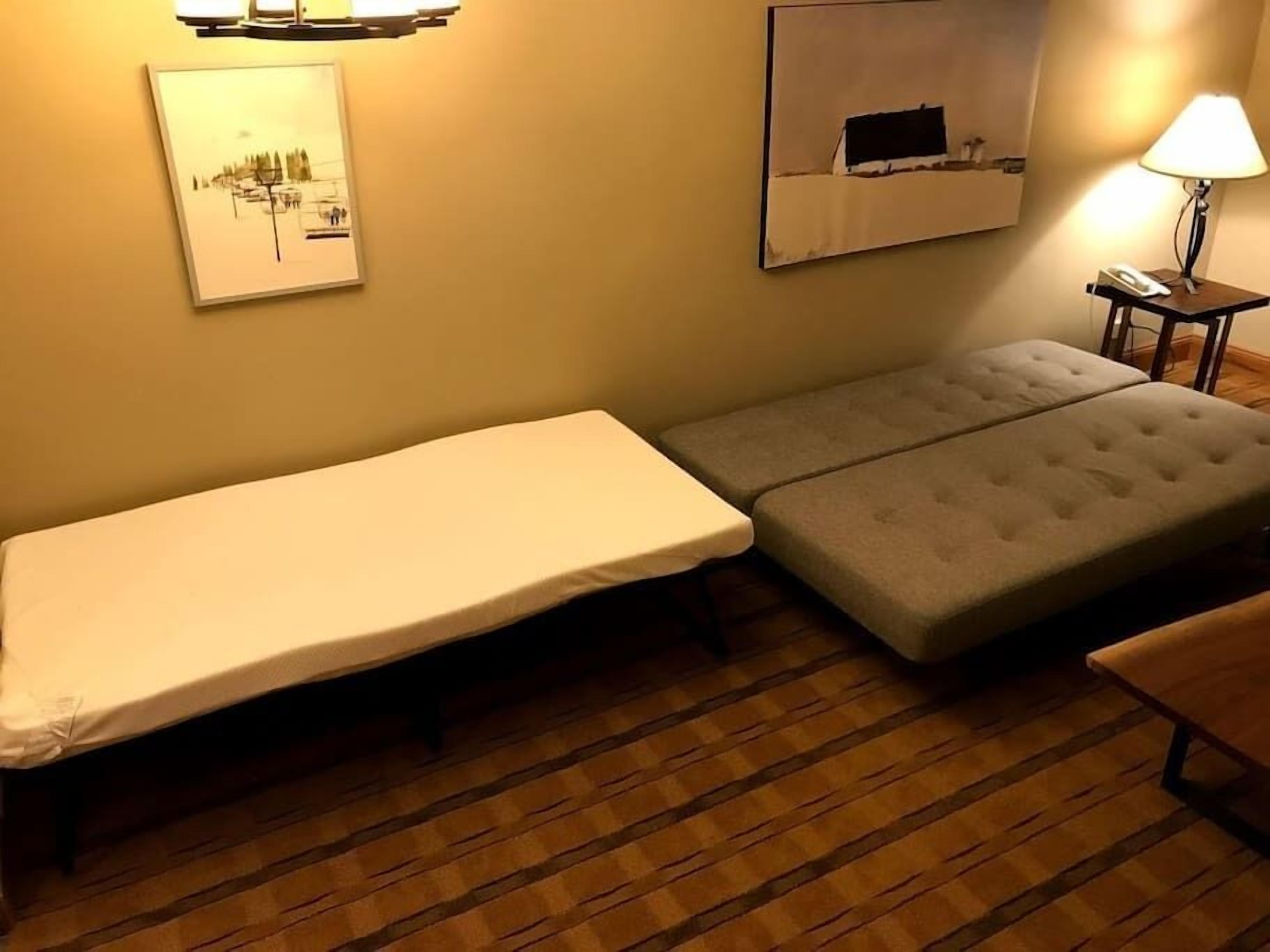 Una habitación pequeña con una cama individual, un banco, una lámpara y un cuadro en la pared.