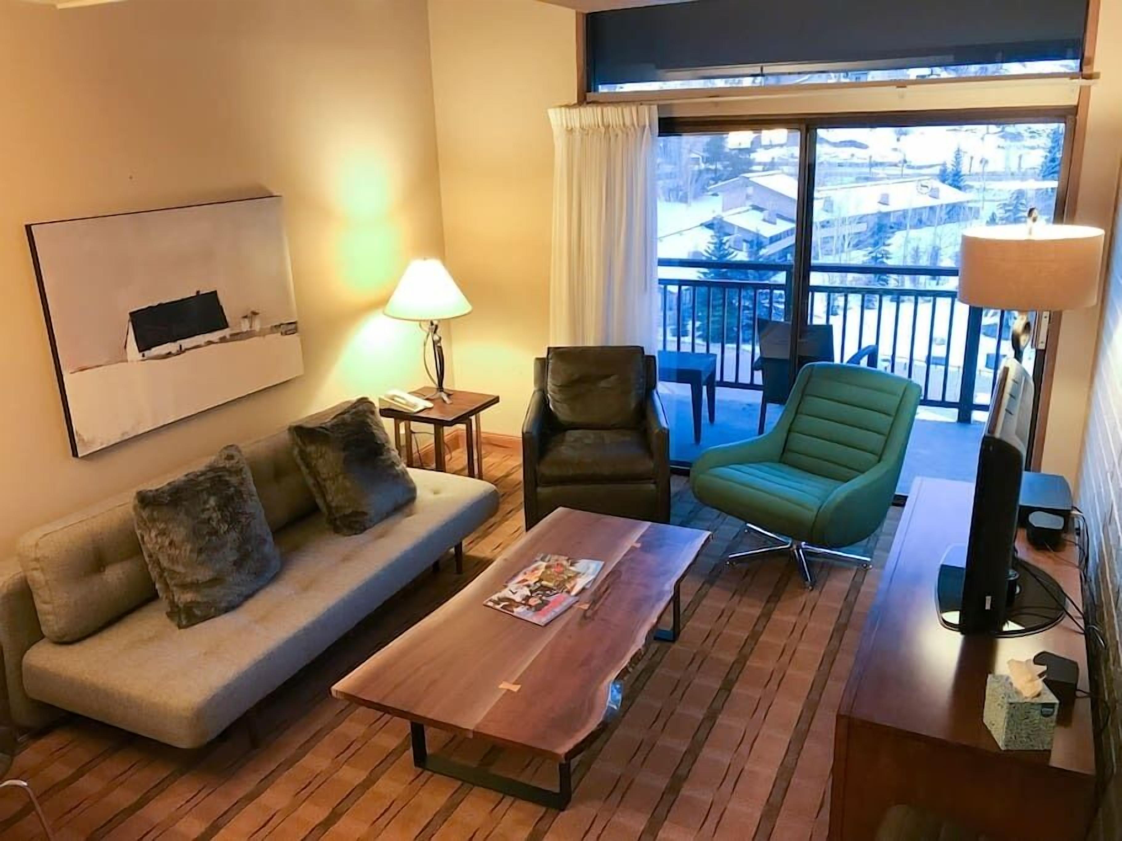 Een gezellige woonkamer met een bank, een groene fauteuil, een houten salontafel en een televisie. Door het raam is een balkon met uitzicht op gebouwen te zien.