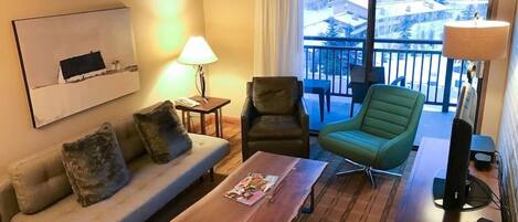 Een gezellige woonkamer met een bank, een groene fauteuil, een houten salontafel en een televisie. Door het raam is een balkon met uitzicht op gebouwen te zien.