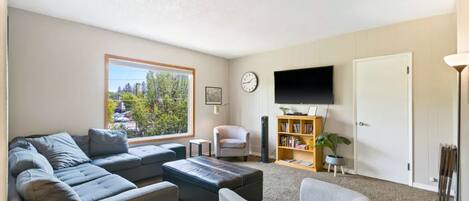 A living room with a grey sofa, a white armchair, a bookshelf, a flat-screen TV, and a window with a view of trees and buildings.